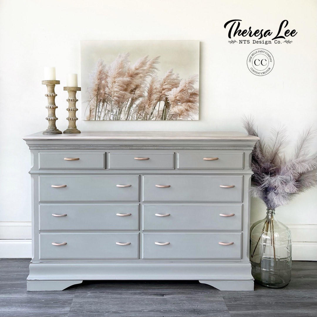 A light gray painted wooden dresser with six drawers and small metallic handles, styled in a room setting with a canvas print, candles, and a glass vase with dried grass.