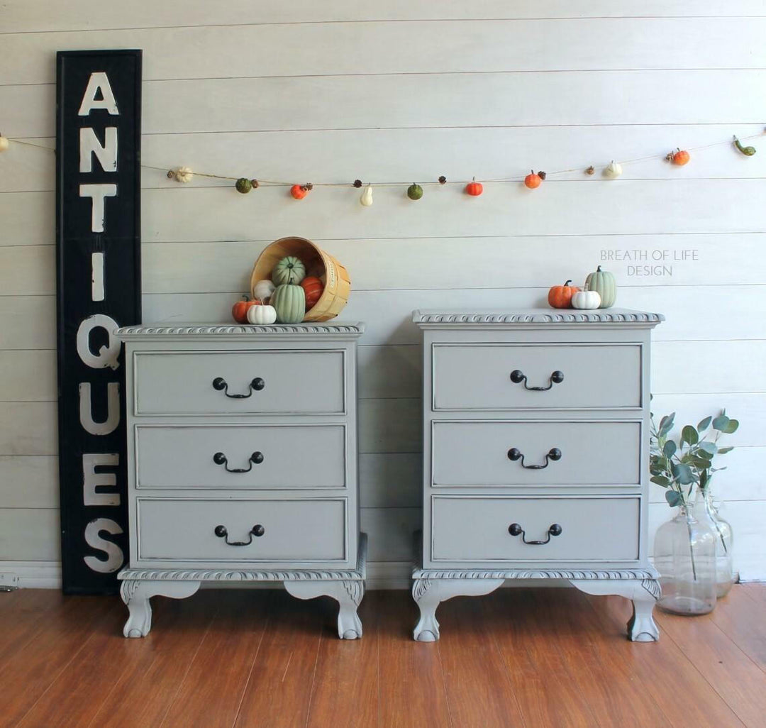 A pair of light gray three-drawer nightstands with black metal handles, styled in a fall-themed room with a black 'ANTIQUE' sign, autumn garland, pumpkins, and a glass vase on a wood floor.