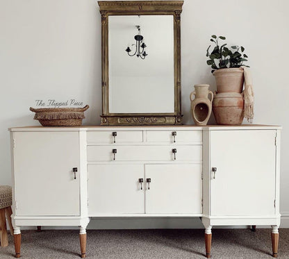 A white wooden sideboard or credenza with dark metal drawer and cabinet pulls, shown in a styled room setting. The piece features a light wood top, four legs with turned wood feet, and is decorated with a large ornate gold-framed mirror, a woven basket, stacked ceramic vases with a plant, and a throw blanket.