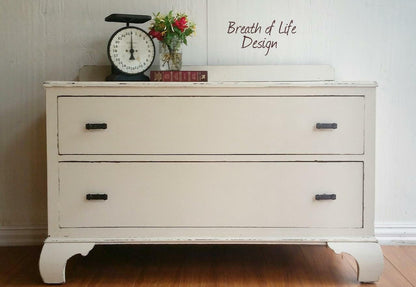 A distressed off-white two-drawer dresser with dark metal handles, styled with a vintage scale, flowers, and books on top, set against a white paneled wall with 'Breath of Life Design' text.