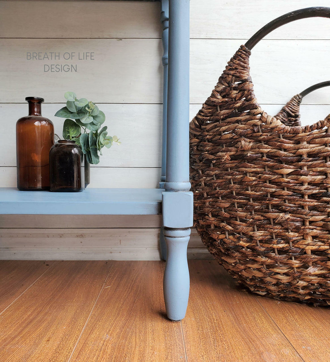 A close-up of a painted wooden table leg in a dusty light blue, positioned next to a large brown woven basket on a wooden floor, with decorative amber glass bottles and a small plant on the tabletop.