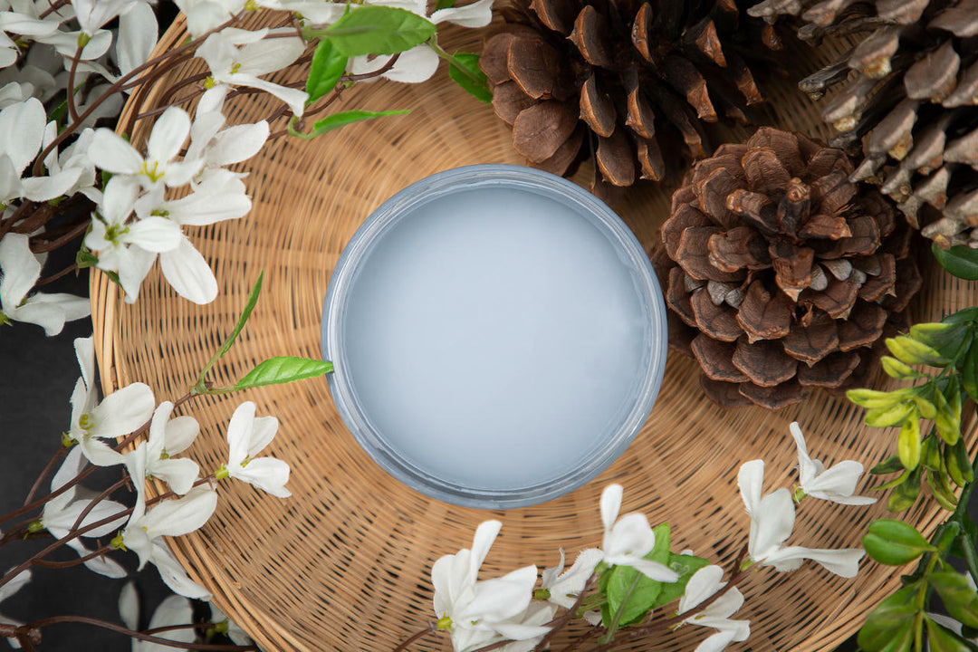 A round glass jar filled with pale blue paint, placed on a woven wicker tray and surrounded by white blossoms, pinecones, and green leaves.