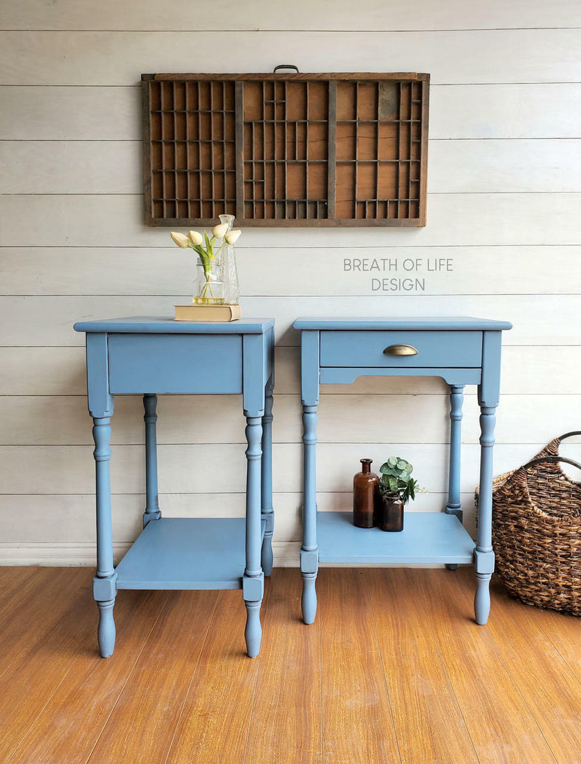 Two matching light blue painted wooden end tables with single drawers and lower shelves, styled in a rustic room setting with a wood floor, white shiplap wall, and decorative items.