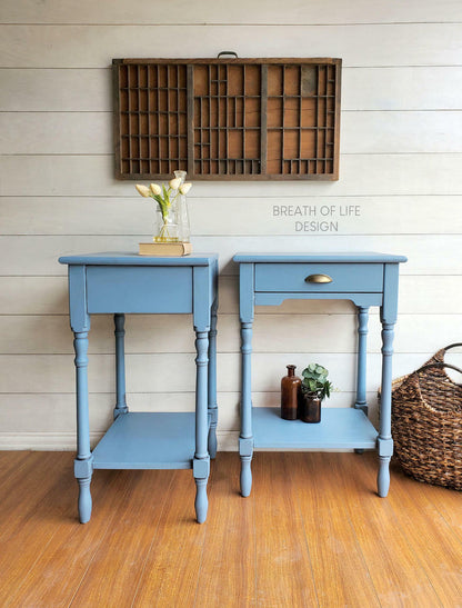 Two matching light blue painted wooden end tables with single drawers and lower shelves, styled in a rustic room setting with a wood floor, white shiplap wall, and decorative items.