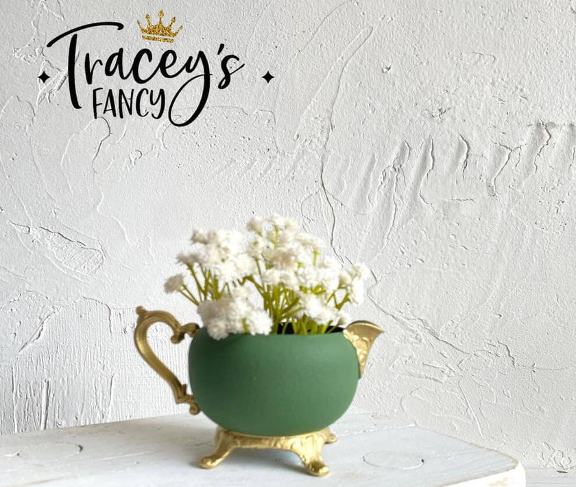 A green ceramic teapot-shaped planter with gold-toned feet and spout, filled with white baby's breath flowers, set against a textured white wall. The brand name 'Tracey's FANCY' with a gold crown logo is displayed in the upper left corner.