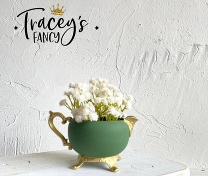A green ceramic teapot-shaped planter with gold-toned feet and spout, filled with white baby's breath flowers, set against a textured white wall. The brand name 'Tracey's FANCY' with a gold crown logo is displayed in the upper left corner.