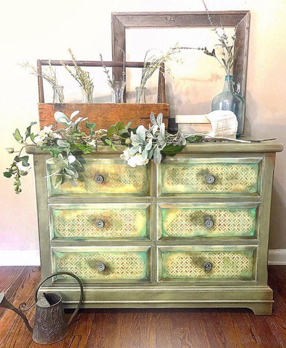 A distressed sage green and yellow painted wooden dresser with six drawers, styled with a rustic wooden crate, white flowers, a blue glass bottle, and a large mirror on a wood floor against a beige wall.