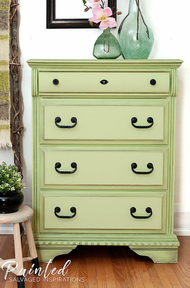 A sage green painted wooden dresser with four drawers and black metal handles, styled in a home setting with decorative vases on top, a small stool, and a wood floor.