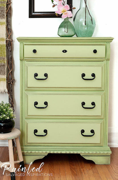 A sage green painted wooden dresser with four drawers and black metal handles, styled in a home setting with decorative vases on top, a small stool, and a wood floor.