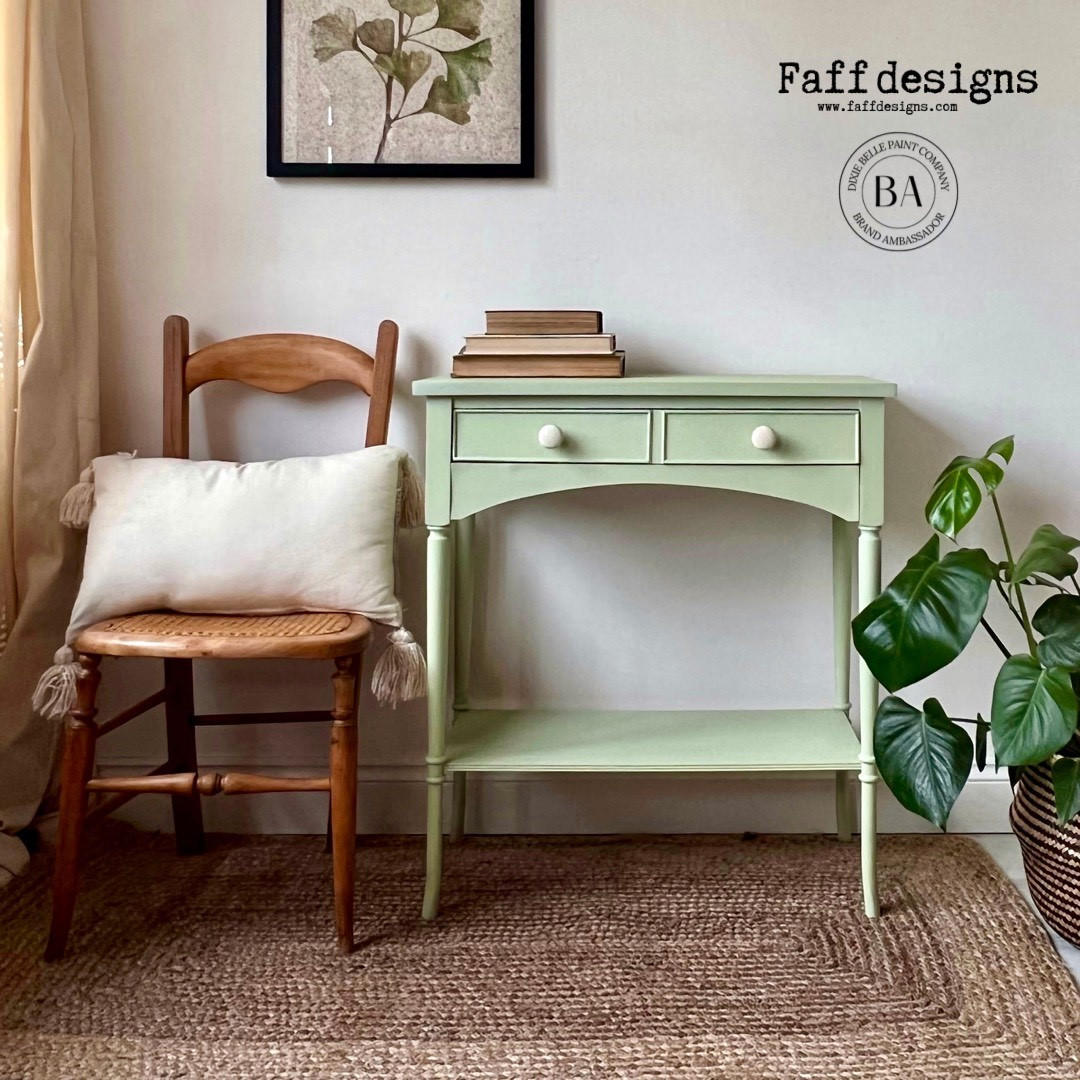 A sage green console table with two drawers and a lower shelf, styled in a cozy room next to a wooden chair with a cream pillow and a potted green plant. The wall features a framed botanical print and the text 'Faff designs' with a BA logo.