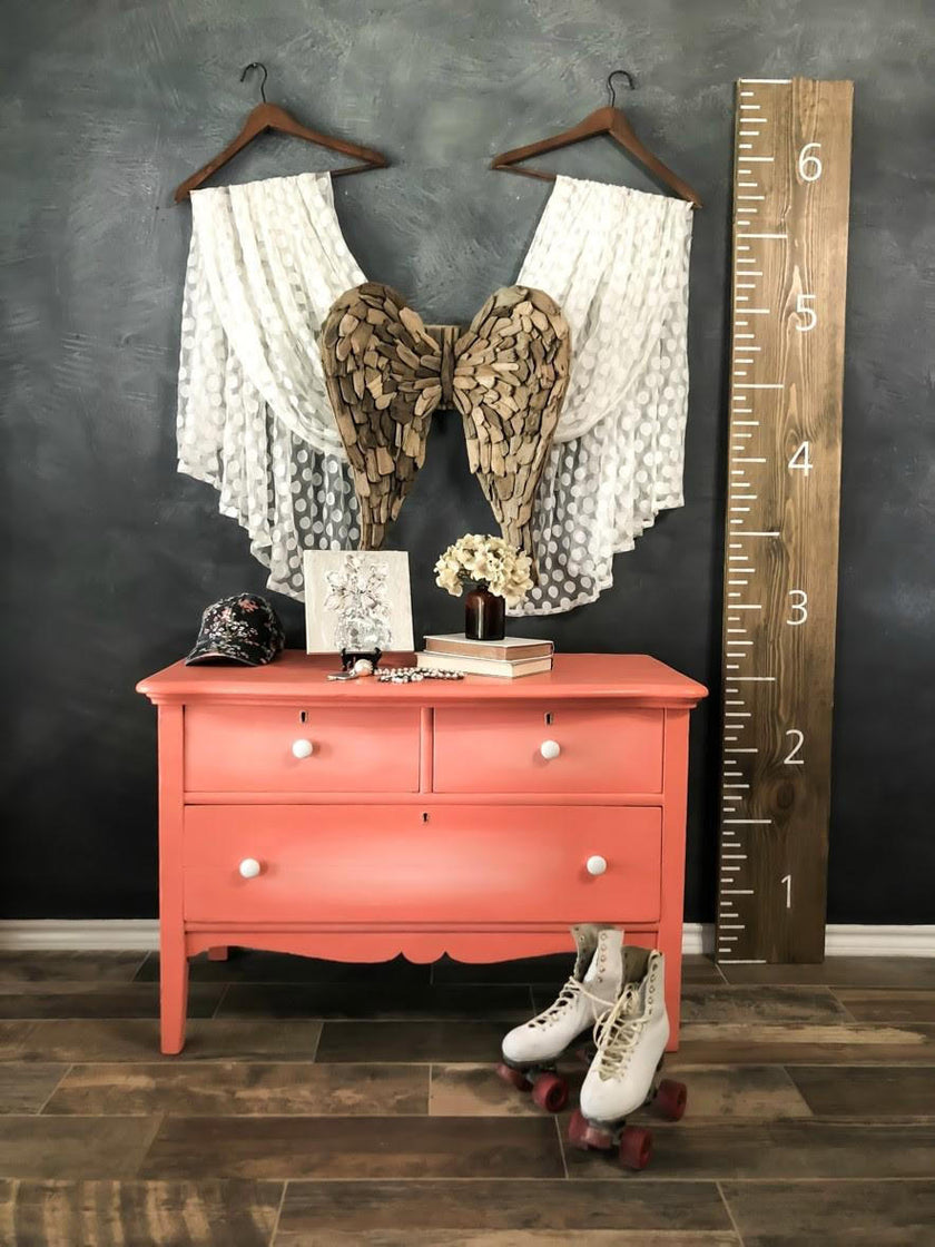 A coral pink painted dresser styled in a rustic room setting, featuring a dark gray chalkboard wall, a wooden floor, a tall wooden height ruler, and white lace-up roller skates on the floor. Decorative items including a lace angel wing, a wooden heart, and lace garments hang on wooden hangers above the dresser.