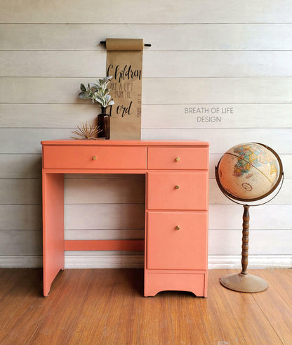 A coral pink wooden writing desk with three drawers and small brass knobs, styled in a room with a white shiplap wall, wood floor, a hanging scroll, and a vintage globe.