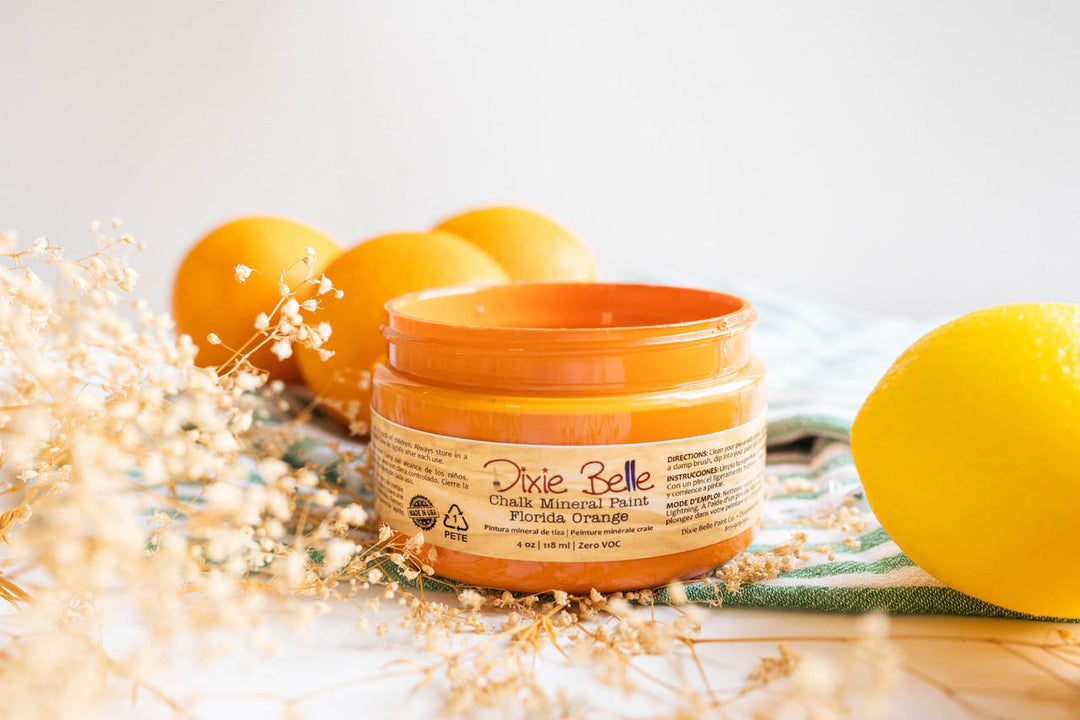 A jar of Dixie Belle Florida Orange Chalk Paint, styled with fresh oranges and a lemon on a striped towel with dried baby's breath flowers.