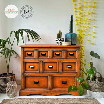 An orange-brown wooden 9-drawer chest of drawers styled in a home interior setting with potted plants, a cactus, and decorative hanging garlands.