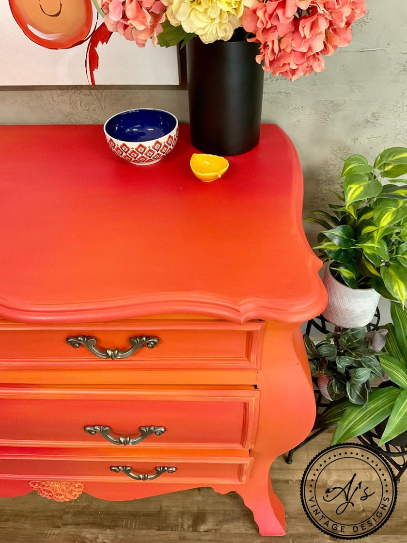 A bright orange-red painted wooden chest of drawers with three drawers and ornate silver handles, styled with a black vase of flowers, a blue and white bowl, and a small yellow bowl on top. A potted green plant sits to the right, and a decorative circular watermark is visible in the bottom right corner.