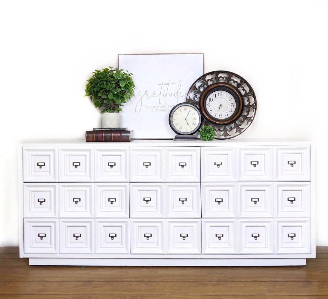 A white multi-drawer storage cabinet with small drawer pulls, styled against a white wall on a wood floor with a potted plant, books, a framed print, a small clock, and a decorative round wall clock on top.