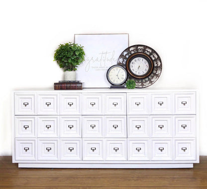 A white multi-drawer storage cabinet with small drawer pulls, styled against a white wall on a wood floor with a potted plant, books, a framed print, a small clock, and a decorative round wall clock on top.