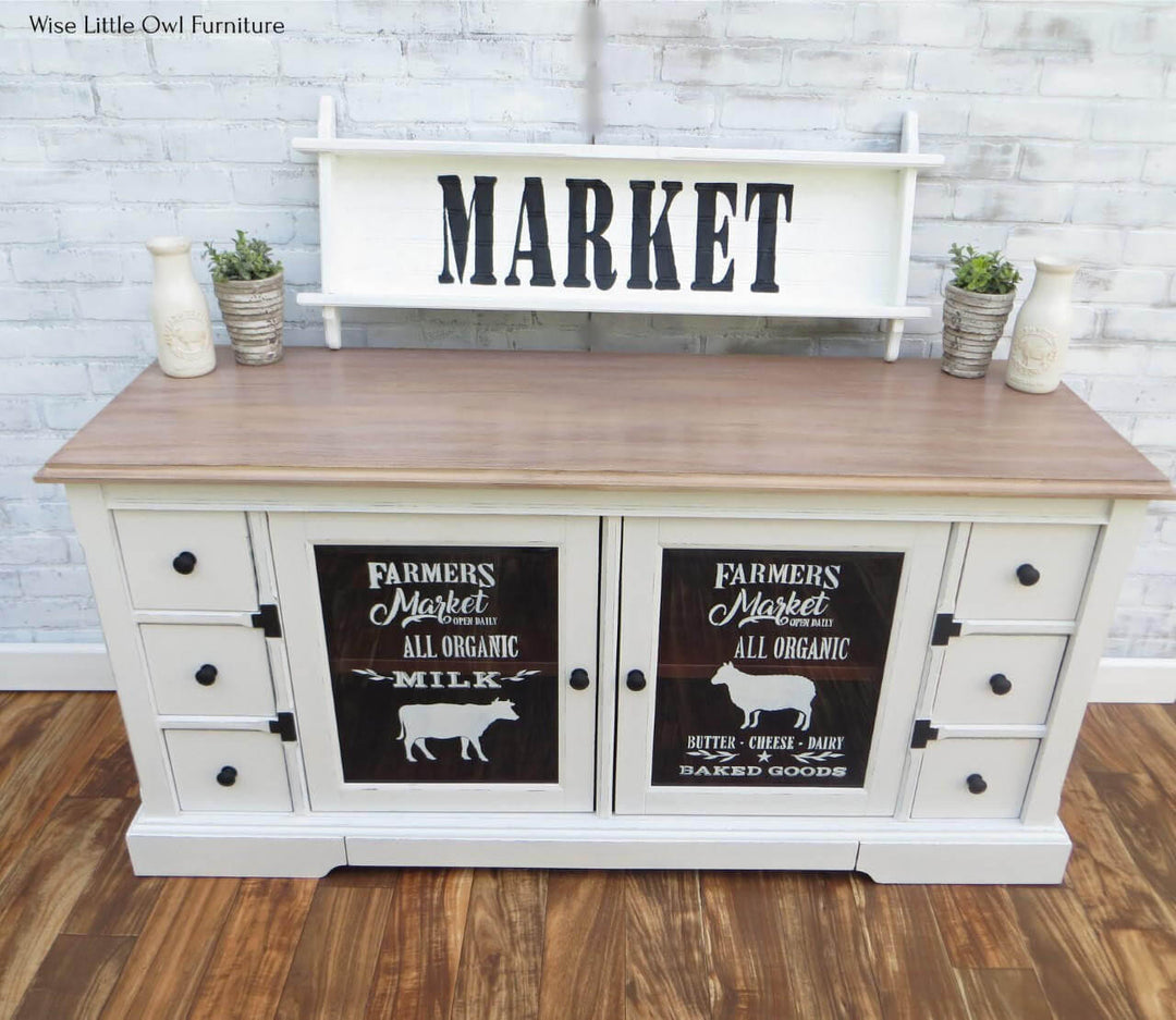 A white farmhouse-style sideboard cabinet with a light wood top, decorative black-and-white sign panels on the doors, and a matching white shelf mounted above. The cabinet is styled in a rustic room with a white brick wall and wood floor.