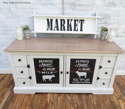 A white farmhouse-style sideboard cabinet with a light wood top, decorative black-and-white sign panels on the doors, and a matching white shelf mounted above. The cabinet is styled in a rustic room with a white brick wall and wood floor.