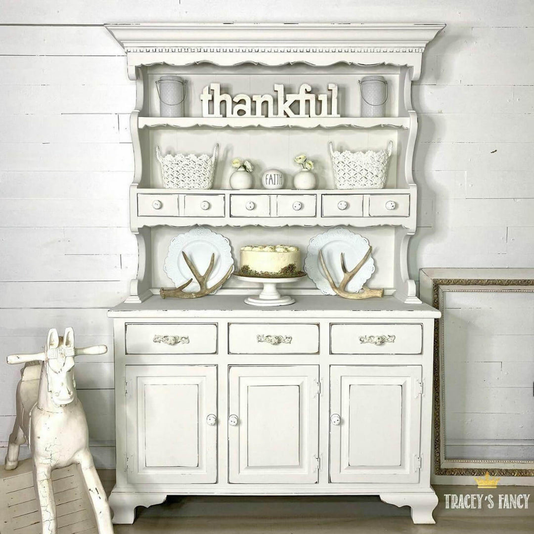 A distressed white shabby-chic hutch and sideboard cabinet styled with white ceramic dishes, baskets, and a small cake stand, set against a white shiplap wall with a wooden rocking horse on the left.