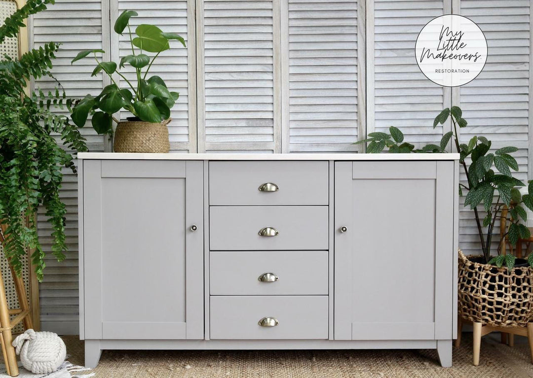A light gray painted wooden sideboard or credenza with three central drawers and two outer doors, styled in a room setting with potted green plants, a woven basket, and white shuttered windows in the background. A circular logo for 'My Little Makeup' is visible in the top right corner.