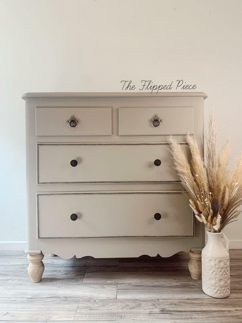A three-drawer wooden dresser in a light beige or greige finish with dark metal knobs, styled in a room setting next to a vase of dried pampas grass. The dresser has a scalloped lower front and turned legs.