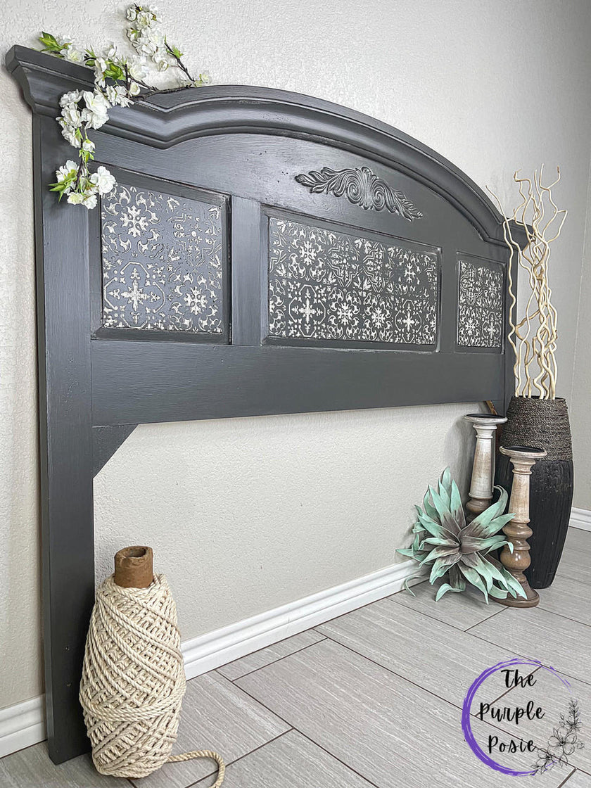 A dark charcoal gray upholstered headboard with three decorative floral panels, styled in a bedroom setting with a large spool of twine, a vase with dried branches, and a plant on a light wood floor.
