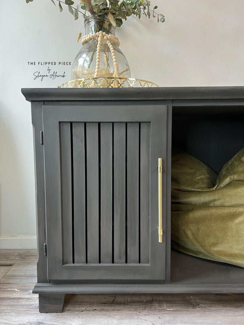 A dark gray wooden storage cabinet with a slatted door and a brass handle, shown in a styled room setting with a green bean bag inside and a glass vase on top.
