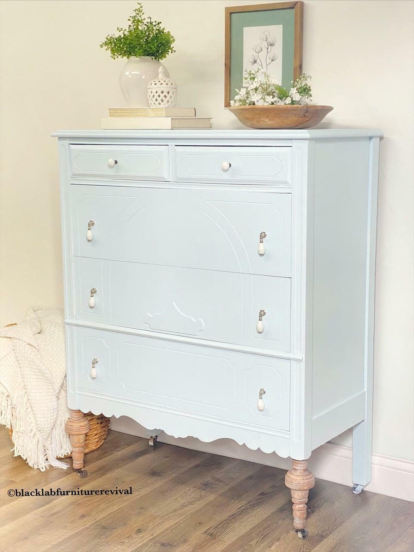 A light blue painted wooden chest of drawers with distressed brown turned legs and small silver knobs, styled in a room setting with home decor items on top.