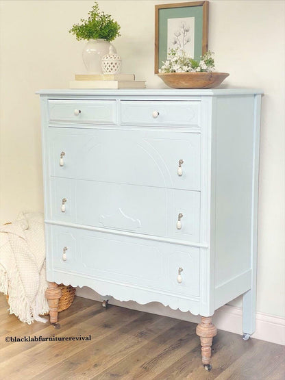 A light blue painted wooden chest of drawers with distressed brown turned legs and small silver knobs, styled in a room setting with home decor items on top.