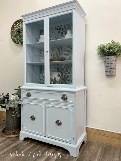 A tall, light blue painted hutch cabinet with two glass-front upper doors, open shelves, a single drawer, and two lower doors, styled in a home interior setting.