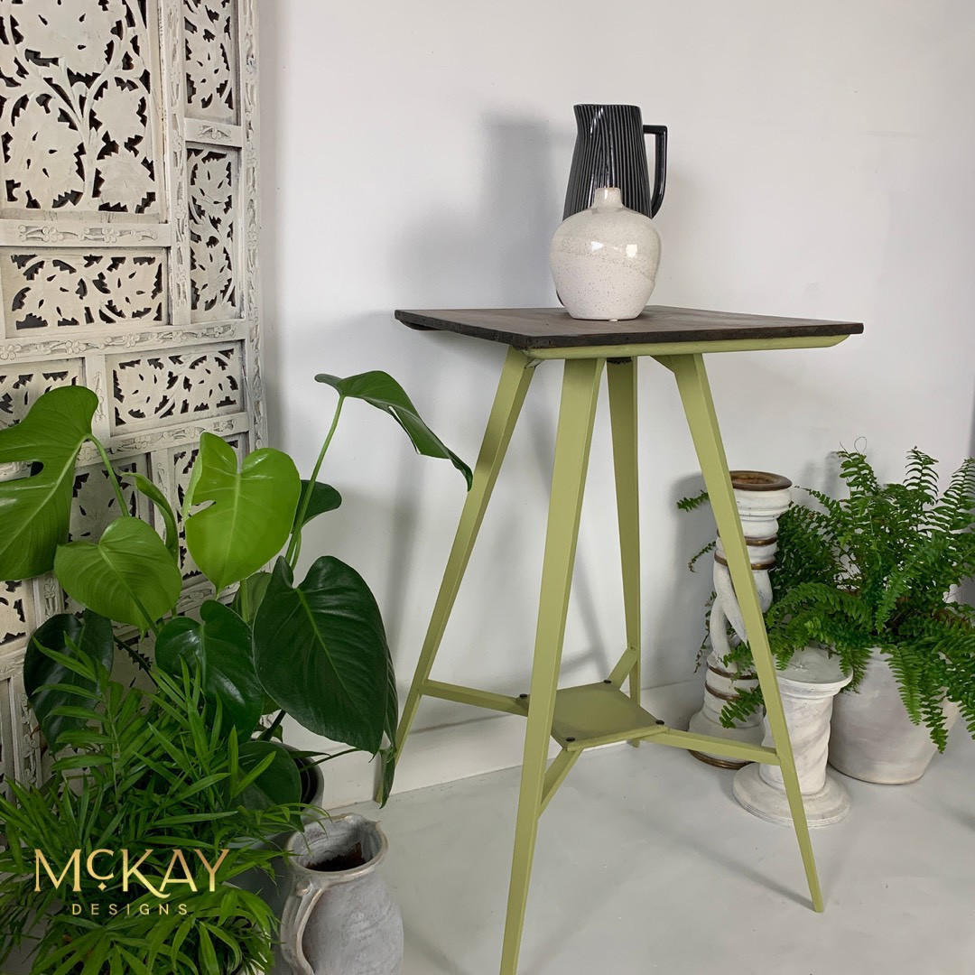 A three-legged accent table with a dark wood top and a sage green painted metal frame, styled with a white ceramic pitcher on top. The table is set in a bright room with potted green plants, a white carved wooden screen, and decorative pedestals.