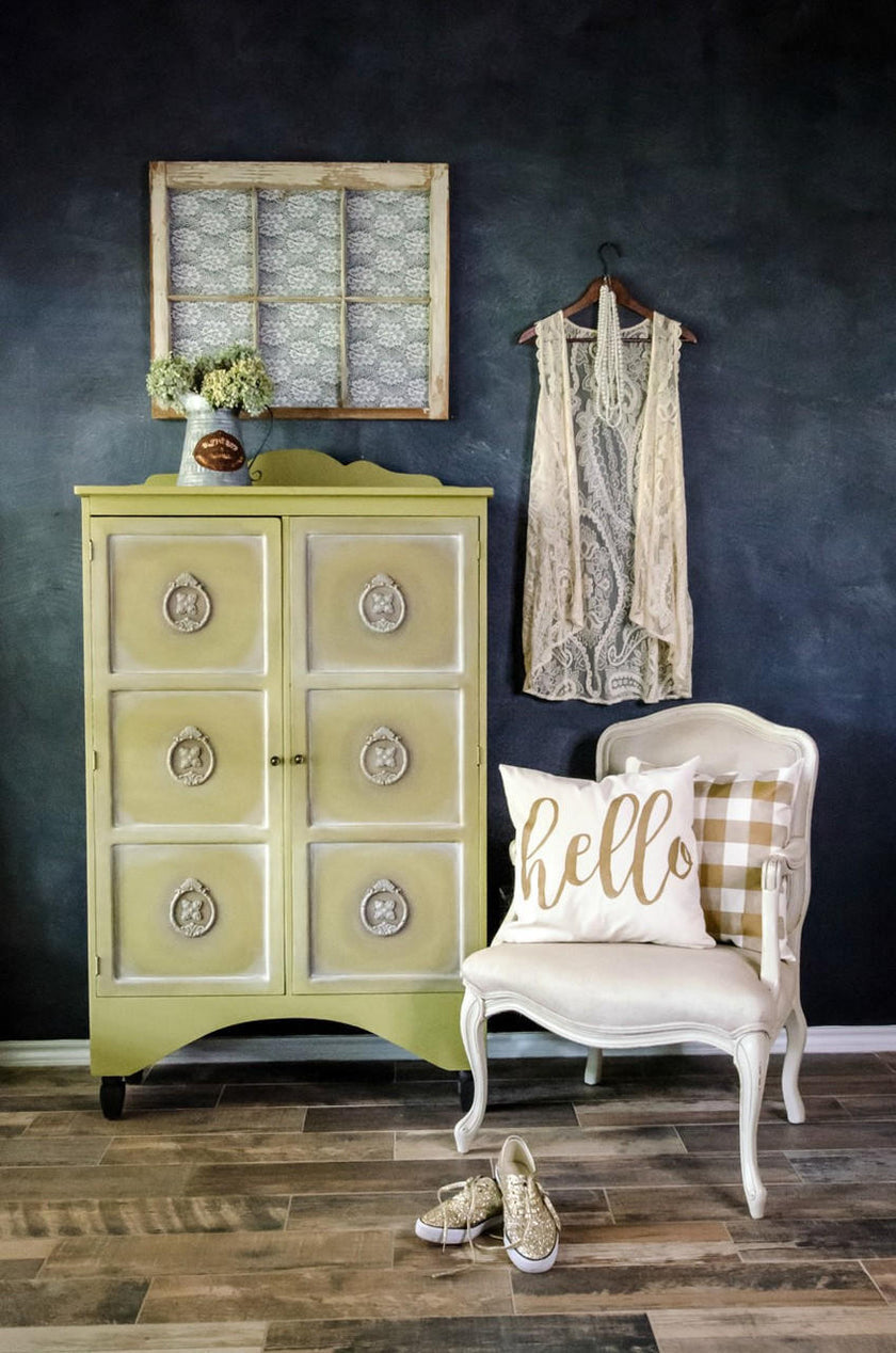 A styled interior scene with a pale yellow-green and white vintage-style cabinet against a dark wall, a cream chair with a 'hello' pillow, a lace garment hanging on a hanger, and shoes on the wood floor.