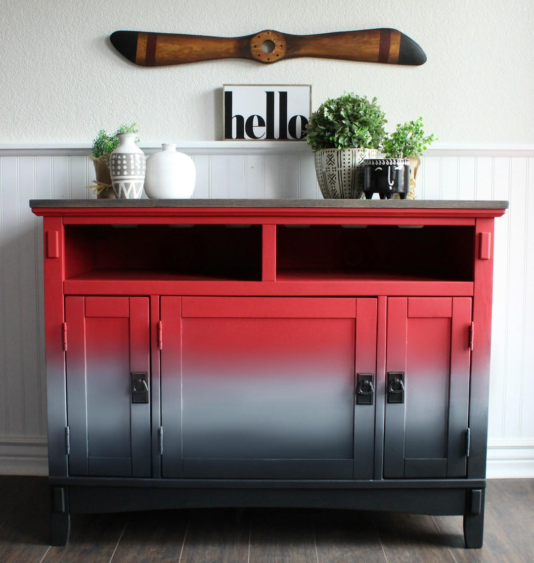 A wooden sideboard cabinet with a red-to-black ombre gradient finish, styled with home decor items on top against a white paneled wall.