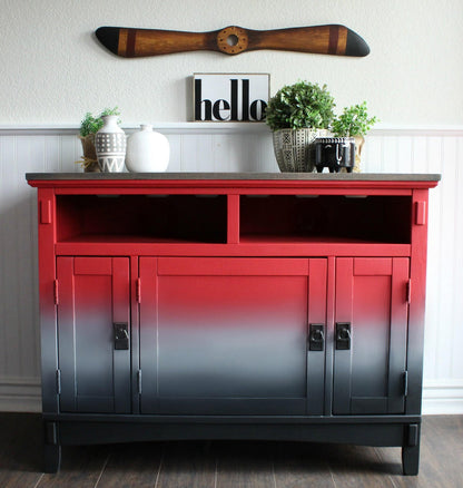 A wooden sideboard cabinet with a red-to-black ombre gradient finish, styled with home decor items on top against a white paneled wall.