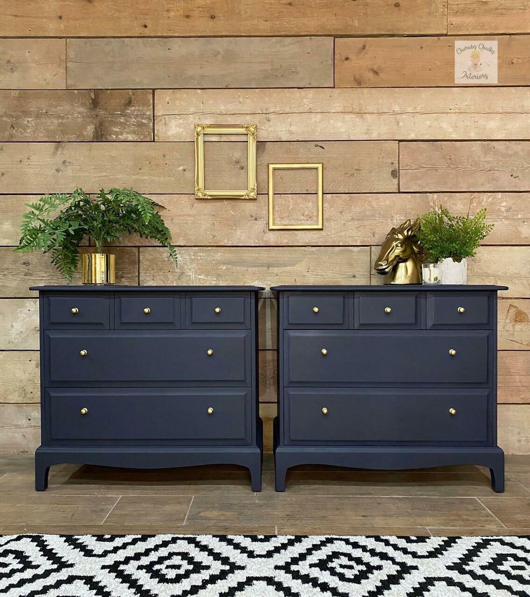 A pair of navy blue wooden chest of drawers with brass knobs, styled against a rustic wood plank wall with decorative plants and picture frames.