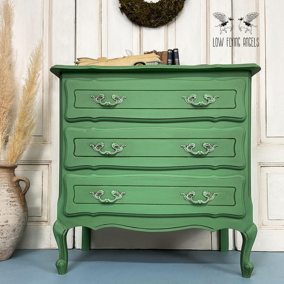 A green painted wooden chest of drawers with four drawers and ornate silver handles, styled in a room setting with a vase of pampas grass, a wreath, and books on top. The Low Flying Angels brand logo is visible in the top right corner.