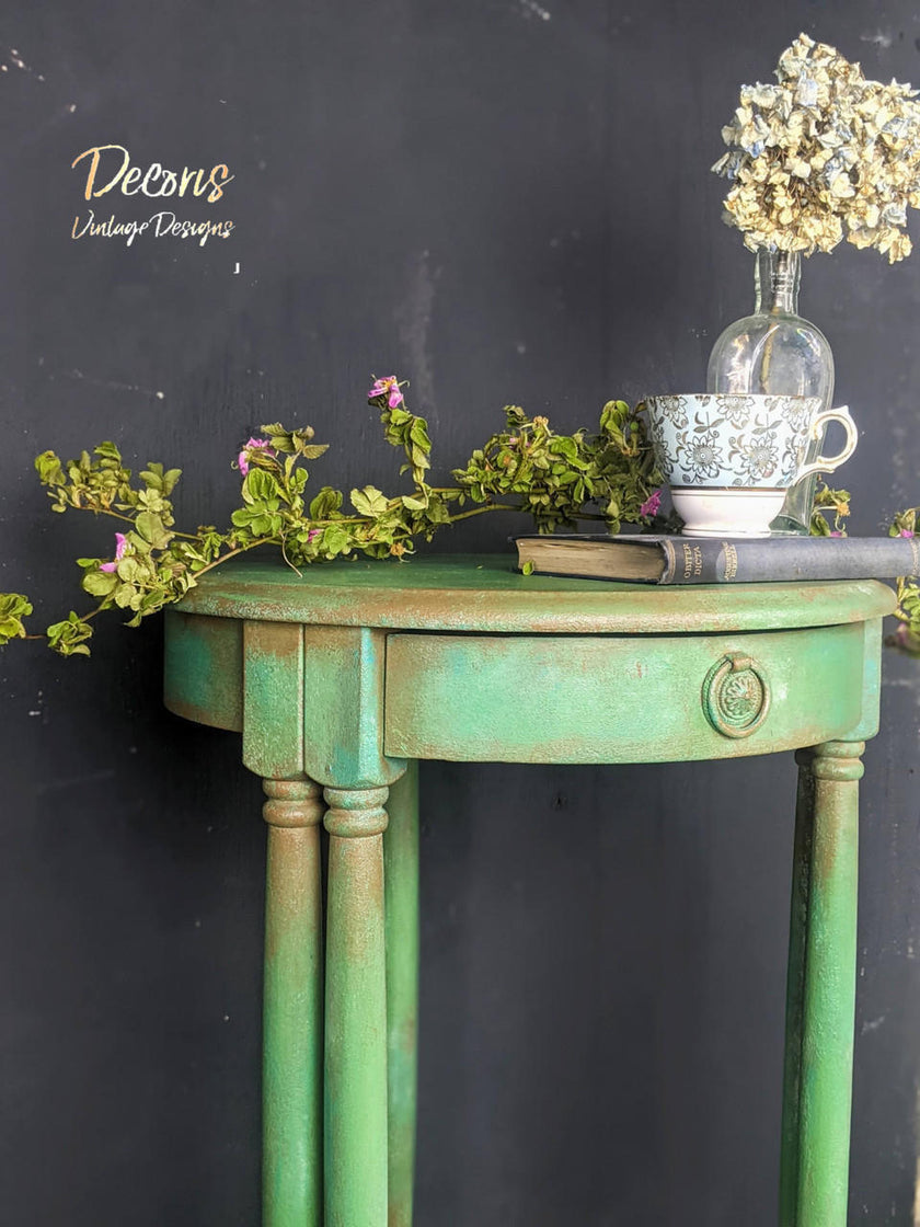A distressed sage green side table with a single drawer and turned legs, styled with a teacup, vase of dried flowers, and greenery against a dark chalkboard background. 'Deons Vintage Designs' logo is visible in the upper left.