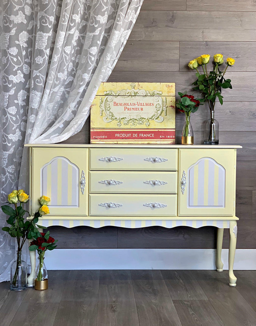 A pale yellow and white painted wooden sideboard or buffet cabinet with three drawers and two arched door panels, styled against a wood-paneled wall with curtains and flower vases.