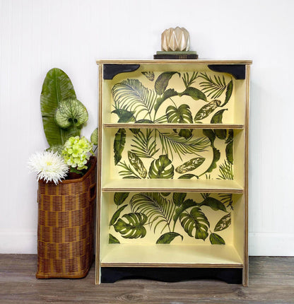 A three-tier wooden bookcase with a tropical leaf pattern on the shelves, styled next to a wicker basket with flowers against a white paneled wall.
