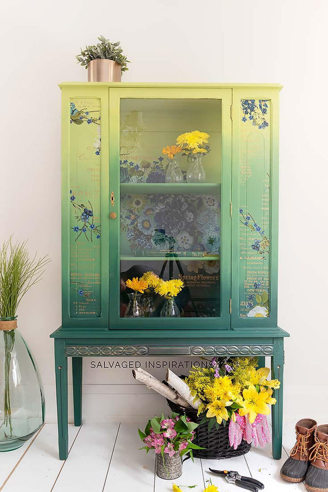 A vintage-style painted wooden cabinet with a green-to-yellow gradient and floral decals, styled in a bright room with flowers, a plant, and shoes on a white floor.