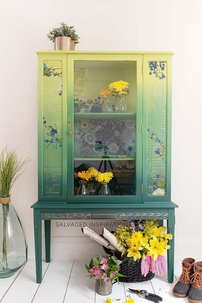 A vintage-style painted wooden cabinet with a green-to-yellow gradient and floral decals, styled in a bright room with flowers, a plant, and shoes on a white floor.