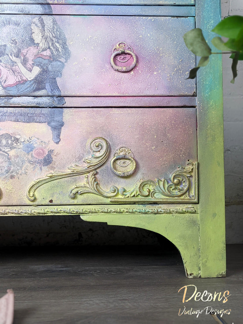 A close-up of a vintage-style painted dresser with distressed pastel colors, floral artwork, ornate gold drawer pulls, and a green-painted leg, shown indoors with a plant leaf entering from the right.