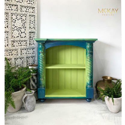 A small painted wooden cabinet or shelf with a bright lime green interior and turquoise-to-blue turned legs, styled against a white wall with decorative plants and vases. The brand name 'MCKAY DESIGNS' appears in the top right corner.