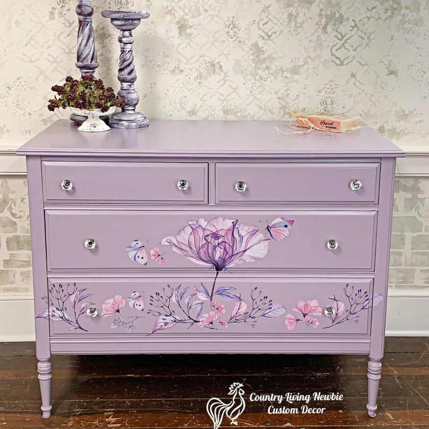 A lavender purple painted wooden dresser with floral and butterfly hand-painted details, styled in a home setting with decorative candlesticks and a small dish on top.