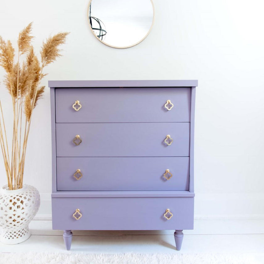 A lavender purple three-drawer chest of drawers with gold quatrefoil and ring pulls, styled in a bright room next to a white vase with dried pampas grass.