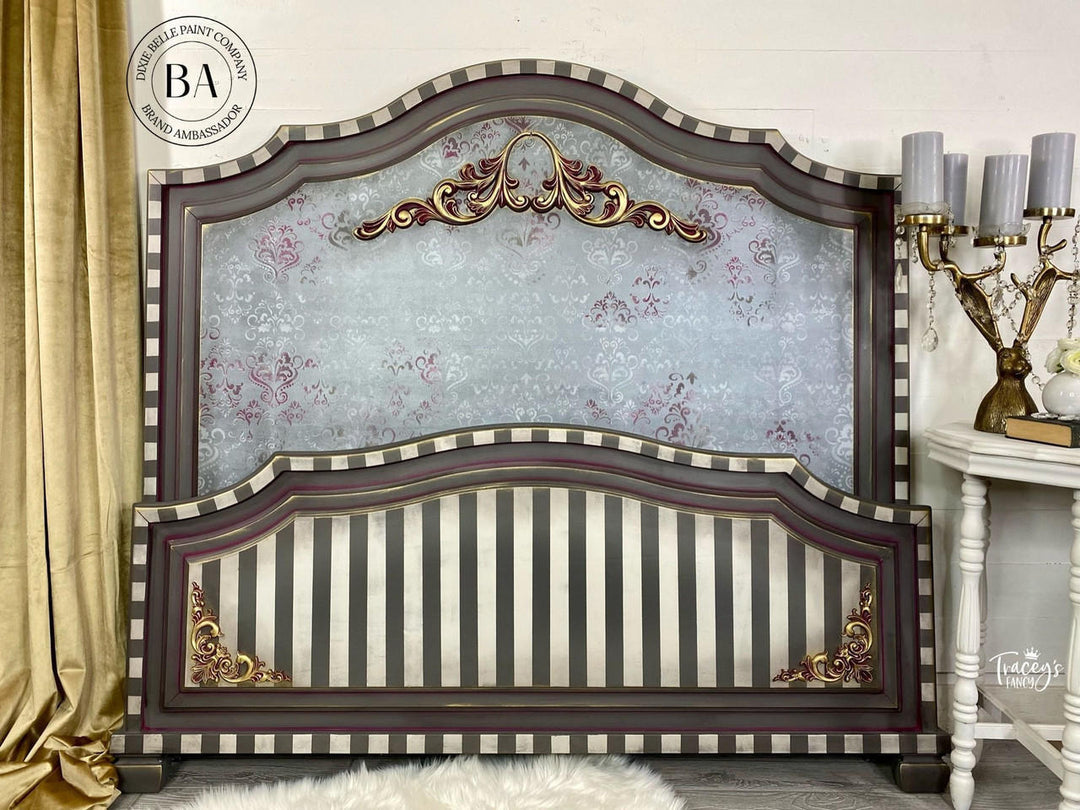 An ornate wooden headboard with a dark brown frame, striped panels, and gold accents, set against a light gray wall with a gold curtain and a side table visible.