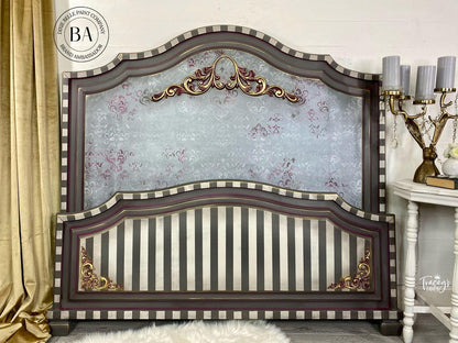 An ornate wooden headboard with a dark brown frame, striped panels, and gold accents, set against a light gray wall with a gold curtain and a side table visible.
