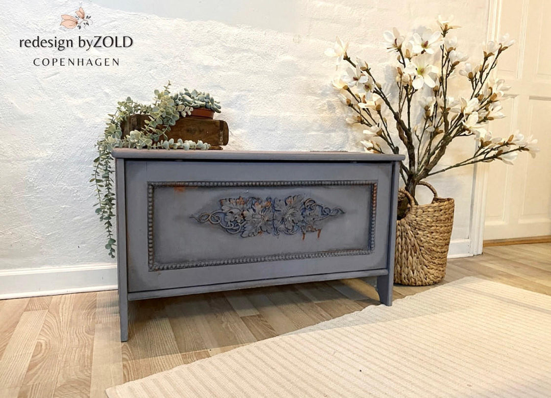 A painted wooden storage chest or bench with a distressed gray finish and ornate floral detailing on the front panel, styled in a room setting with a textured white wall, light wood floor, and decorative plants.