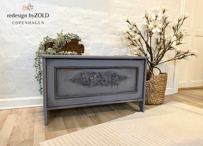 A painted wooden storage chest or bench with a distressed gray finish and ornate floral detailing on the front panel, styled in a room setting with a textured white wall, light wood floor, and decorative plants.